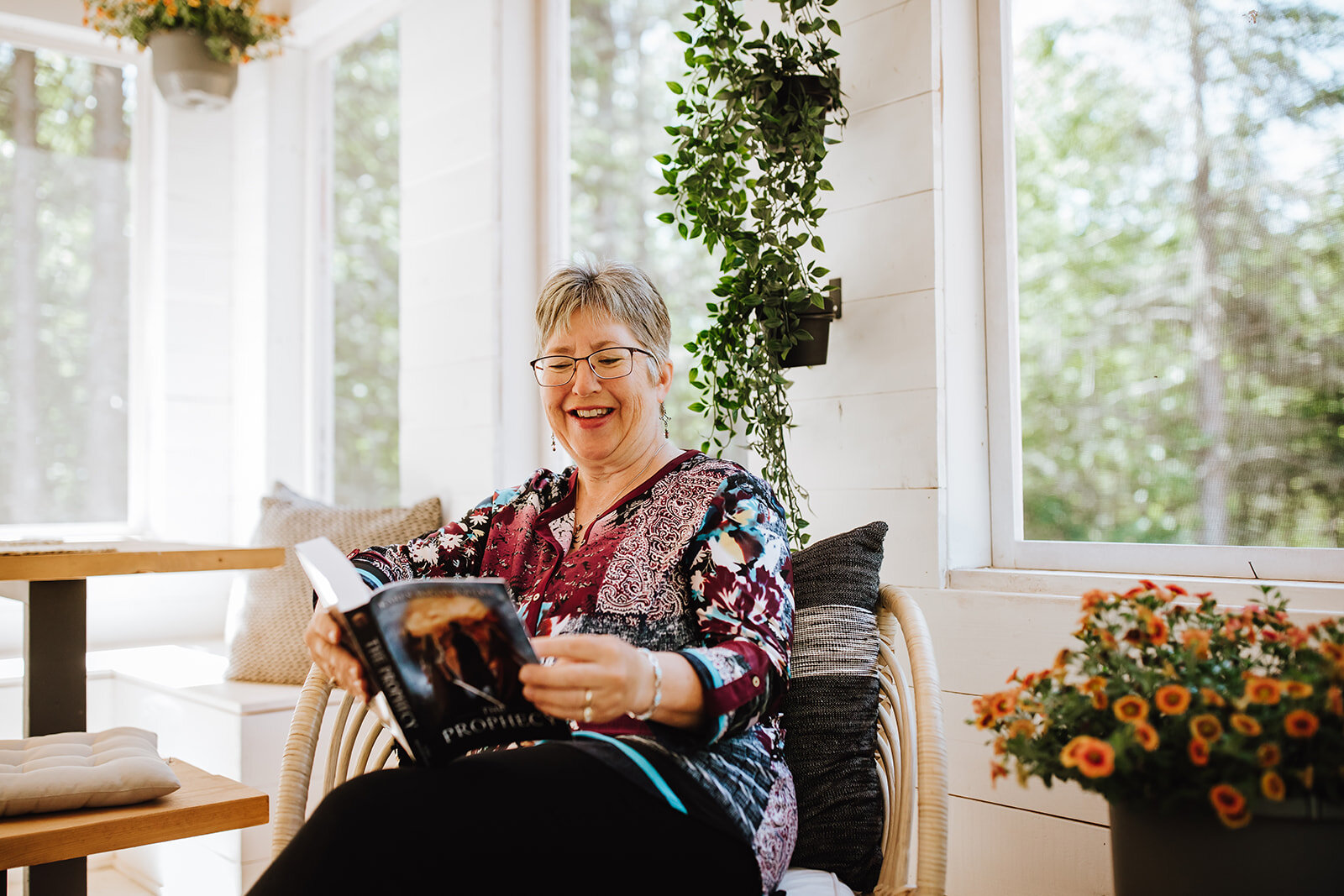 The Author Karen MacLeod-Wilkie sits in a cushioned wicker chair smiling while reading The Prophecy book. She is surrounded by multiple plants and large windows brightly lighting the space.